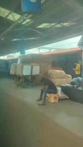 A handcart loaded with burlap sacks and white boxes sits on a paved surface next to a train platform. In the background, people are visible near water coolers and a train. The scene is under a covered structure with a corrugated metal roof.