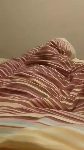The video shows a person under a striped blanket on a bed. The person is moving and appears to be talking, but their face is not visible. The room is dimly lit, and the focus is on the blanket and the movement beneath it.