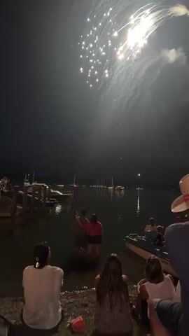 The video shows a fireworks display at night over a body of water. Several people are gathered on the shore watching the fireworks. Some people are standing in the water. There are boats docked on the left side of the frame. The fireworks are bright and scattered across the sky. The scene is dimly lit, with the fireworks providing the main light source.