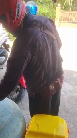 The video begins with a close-up of a person wearing a red helmet and a dark jacket, standing next to a red motorcycle. The motorcycle has a license plate that reads "W 2990 MAM". The person appears to be adjusting something on the motorcycle. The camera then pans slightly to reveal more of the environment. There are other people in the background, and what appears to be a street or paved area. In the foreground, there is a yellow object, possibly a storage container, and a large, round, grey object. The lighting suggests it is daytime. The camera movement is shaky, indicating it was likely handheld. The video captures a brief moment of interaction with the motorcycle.