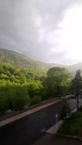 The video captures a view of a lush, green forested hillside under a dramatic, overcast sky. The sun is breaking through the clouds in the distance, creating a bright, hazy glow over the landscape. A paved road runs horizontally through the lower part of the frame, with a few parked cars visible. A small, triangular warning sign is positioned on the side of the road. Tall trees dominate the middle ground, with a particularly large, rounded tree standing out. In the foreground, on the right side, a portion of a brick building is visible, with two satellite dishes attached to its exterior. The scene evokes a sense of moody natural beauty, perhaps during or just after a rain shower, given the softness of the light and the slightly damp appearance of the road. The time of day appears to be late afternoon or early evening, judging by the quality of the sunlight.