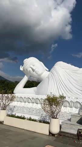 The video pans across a large white statue of a reclining Buddha. The statue is intricately detailed, with folds in the robe and a serene expression on the Buddha's face. The Buddha lies on a decorative base with lotus flower motifs. Two potted plants with green foliage flank the statue on the right side, and another plant is visible on the left. In the foreground, there's a planter box with pink flowers and a sign that reads "HOLY AREA Area Suci." The background features a clear blue sky with some white clouds and lush green trees on a distant hill. The lighting suggests it is daytime, likely with bright sunlight.
