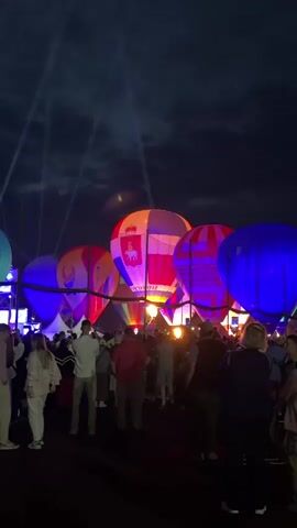 The video shows a night scene with several hot air balloons illuminated by spotlights and internal lighting. The balloons are of various colors, including blue, purple, pink, orange, and red, and some have patterns or logos on them. A crowd of people is gathered in the foreground, facing the balloons and appearing to watch a show or event. The sky is dark, suggesting it is nighttime, and there are light beams cutting through the darkness. The atmosphere seems festive.