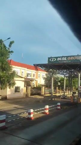 A security camera captures a view of a toll plaza. A large green and white sign with the words "DLGL GROUP" is visible at the top. The plaza has several toll booths, some with attendant booths. The road has barriers and lane dividers. A building with a red roof and light-colored walls is behind the toll plaza. The sky is blue with some clouds. A person in a blue shirt and dark pants is standing near a bench.