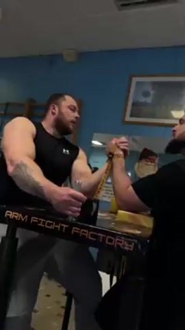 Two men are engaged in an arm wrestling match. The man on the left is wearing a black tank top and gray sweatpants, and has a tattoo on his left arm. He is seated on a stool. The man on the right is wearing a black t-shirt and is seated. Both men have beards. They are at an "Arm Fight Factory" table. The background includes blue walls, a framed picture, a mirror, and gymnastic bars. The arm wrestling match appears to be in progress with both men exerting effort.