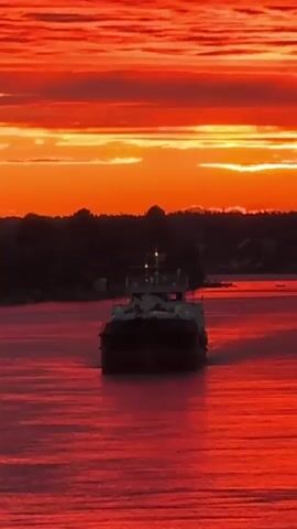 A barge sails on a river at sunset. The sky is a fiery orange and red, with clouds stretching across the horizon. The water reflects the vibrant colors of the sky, creating a mirror-like effect. The barge is dark and silhouetted against the colorful backdrop. Trees and buildings are visible on the distant shore, also silhouetted against the sunset. The scene is peaceful and serene.