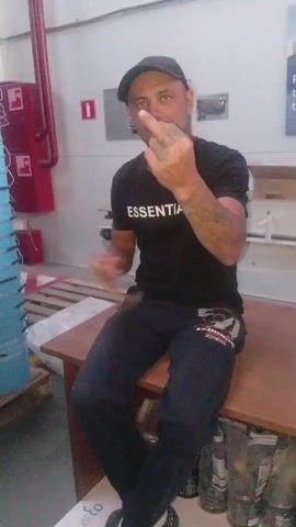 A man wearing a black t-shirt with "ESSENTIALS" on it, dark pants, a baseball cap, and black shoes sits on a wooden table. He is facing the camera and making a middle finger gesture with his right hand. In his left hand, he holds what appears to be a small, elongated object, possibly a snack, which he brings closer to his mouth. The background shows a white wall with some red fire safety equipment on the left and shelving units with various items, including boxes and bottles, on the right. There are also stacks of blue containers and a pallet visible. The lighting is artificial and seems to be overhead.