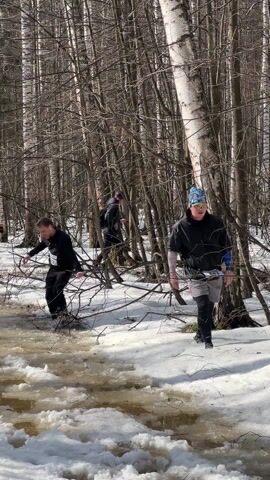 Several people are trudging through a slushy, icy stream in a wooded area on a sunny day. Three people are visible in the foreground. The person on the left wears black clothes, a bib with the number 522, and appears to be splashing through the water. The person on the right is closer to the camera, wearing a black jacket, gray shorts, patterned headwear, and dark leggings. They are also walking through the slush. A third person in dark clothing is behind them further into the woods. The ground is covered in snow and slush, and the woods have a lot of bare trees.
