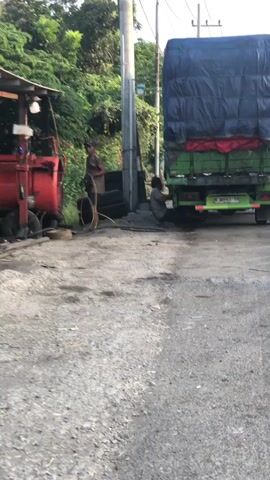 A flat, gravelly surface with a concrete or asphalt area in the foreground. To the left, a red industrial machine with various hoses and attachments is positioned next to a small, open-air structure with a metal roof. Two men are present. One man, wearing a dark shirt and shorts, stands near the red machine, holding a hose. The other man, wearing a light-colored shirt and dark shorts, is kneeling next to the rear of a large, green truck. The truck is covered with a large blue tarp secured with straps, and it appears to be parked on the asphalt or concrete area. Several stacked tires are visible next to the truck. A utility pole stands behind the truck. In the background, there is lush green foliage and a suggestion of more distant trees or hills. The lighting indicates it is daytime, possibly late afternoon, with soft shadows. The overall scene depicts what appears to be a roadside repair or maintenance area for a large truck.