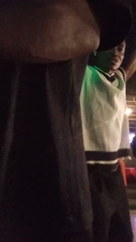 The video captures a nighttime scene in Adonkolo, Nigeria. Initially, a dark, blurry foreground element, possibly a person's leg or arm, occupies the frame. The camera then pans to reveal a person in a white t-shirt and blue pants standing in the distance, holding a phone. They appear to be swaying or moving subtly to music, which is audible in the background. The setting seems to be an outdoor or semi-outdoor area with a visible wooden structure or building in the background, illuminated by artificial lights. The overall atmosphere suggests a casual gathering or event at night. No discernible text is visible.