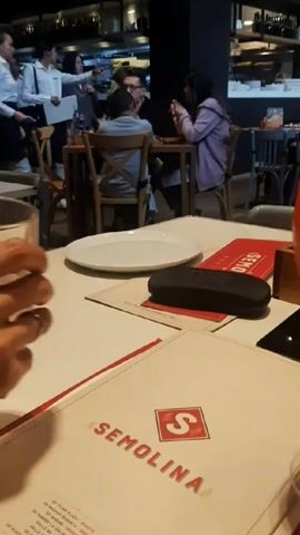 A person is holding a half-full glass and clinking it against a tall glass of orange cocktail in a restaurant.  Other customers are sitting at tables in the background. Staff in white shirts are standing in the kitchen area, visible at the back of the restaurant.