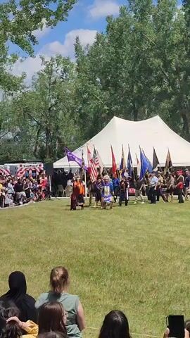 This video captures an outdoor event with a large white tent in the background and trees overhead. A group of people is gathered on a grassy field. In the center of the field, a procession is underway. At the front of the procession, a person in traditional regalia, possibly Native American, holds a purple flag with white symbols. Behind this individual, several other people carry various flags, including the American flag and the Canadian flag. Spectators are visible on the left side of the frame, seated and standing. The overall atmosphere appears to be celebratory or ceremonial.