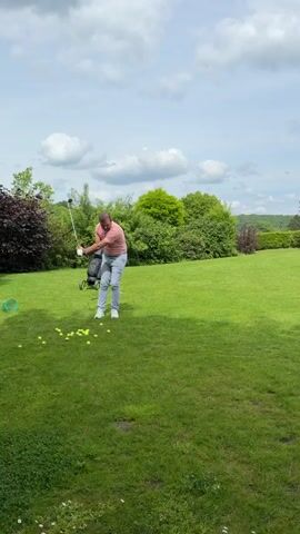 A man wearing a peach-colored shirt and light grey pants takes a golf swing on a lush green lawn. A black golf bag stands beside him. Scattered yellow golf balls are on the ground. The camera then pans to show a golf course with sand traps and flags marking the holes. The sky is partly cloudy.