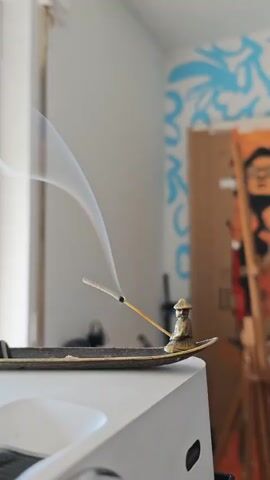 A close-up, static shot captures an incense stick burning in a boat-shaped holder adorned with a small seated figure. Wisps of white smoke curl and drift upwards from the tip of the incense stick. The incense holder rests on a white projector. In the background, a window is visible on the left, with a hint of what appears to be a wall with a blue mural or artwork. The lighting suggests an indoor setting, possibly a room with natural light from the window. The focus is sharp on the incense and smoke, with the background elements softly blurred.