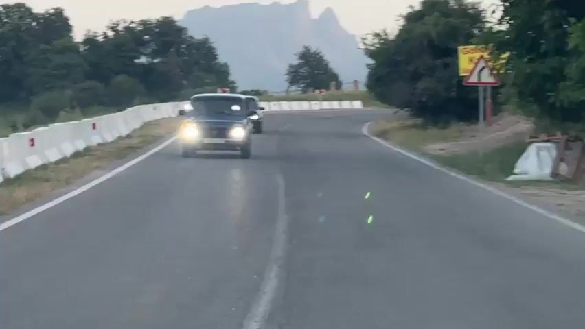 The video shows a winding road with two cars driving on it. The first car, a dark blue SUV, is in the foreground and its headlights are on. The second car, a black sedan, is behind it. The road is bordered by a white concrete barrier on the left and trees and foliage on the right. In the distance, there are mountains. A yellow and red traffic sign is visible on the right side of the road, indicating a sharp left turn. The sky is overcast.