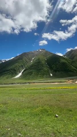 A wide-angle, vertical shot captures a majestic mountain landscape under a bright blue sky dotted with fluffy white clouds. The mountain slopes are covered in lush green vegetation, with patches of snow visible near the peaks. In the foreground, a vibrant green meadow stretches out, interspersed with small yellow wildflowers. A dirt road or track runs along the base of the mountain, separating the meadow from the foothills. The sunlight creates a slight lens flare in the upper right corner of the frame. The overall impression is one of natural beauty and tranquility.