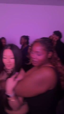 The video captures a lively scene of people dancing at a party under purple lighting. Several individuals are visible, including two women in the foreground who are particularly prominent: one with long dark hair, wearing a leopard-print top, and the other with reddish-brown curly hair in a black top. The woman with dark hair is singing with her mouth open wide. They are dancing closely with their arms around each other. The atmosphere appears energetic and festive, as people wave red cups and sing, with the background featuring other partygoers.