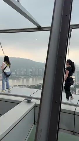 Here's a description of the video:

The video shows a man in a blue shirt and black baseball cap assisting two women wearing safety harnesses on a glass-bottomed observation deck. The man appears to be guiding or instructing the women. The first woman is wearing jeans and a white top with a green accent; she is holding a camera on a selfie stick. The second woman is wearing black athletic clothing. The deck is elevated and offers a panoramic view of a city with many tall buildings, a river or bay, and hills in the background. The sky is overcast, with some sunlight breaking through.
