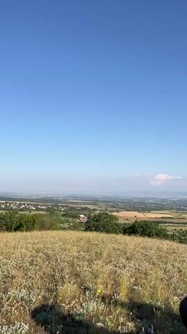 The video pans across a grassy hillside with a quad bike parked in the foreground. The quad bike is black with a large storage box on the back. In the distance, a wide valley stretches out with scattered buildings and fields, leading to a hazy horizon with mountains. The sky is clear and blue with a few scattered clouds. The camera movement is slow and steady, creating a peaceful and scenic view.