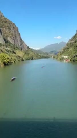 The video shows a wide river or lake, surrounded by steep, rocky, green hills and mountains under a clear blue sky. Two boats are visible on the water, one closer to the left side of the frame and another further in the distance near the center. The water is a greenish-blue color. The scene appears to be captured from a high vantage point, possibly a bridge.