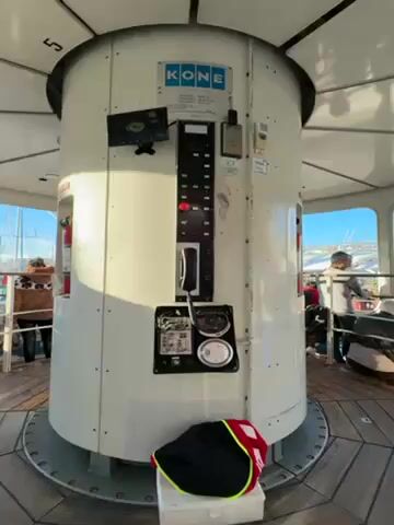 The video shows the interior of a rotating observation tower or structure, possibly a Ferris wheel capsule. A large, cylindrical white control panel is centrally located, labeled "KONE" at the top, and features a control panel with buttons and a phone. A fire extinguisher is recessed into the side of the panel. People are visible through the windows surrounding the structure, looking out at the view. The floor is made of wood planks. The number 5 is visible at the top of the structure. A black bag with a yellow stripe and a white box are at the base of the control panel. The camera rotates slowly around the central control panel.