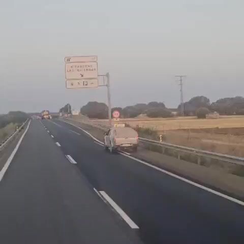 A drone shot follows a vehicle on a highway. The vehicle appears to be a silver pickup truck. The truck is traveling in the right lane, and a yellow combine harvester is visible in the distance, also on the highway. The sky is clear and a pale blue, suggesting it is either sunrise or sunset. The highway is lined with guardrails and vegetation. A sign indicates that Mirabueno and Las Invierno are in the direction of the arrow.
