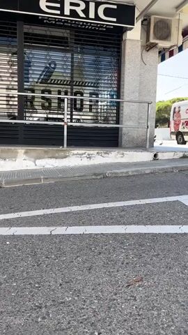 A man on a skateboard attempts a trick on a handrail in front of a commercial building. The buildings appear to be small businesses, including a barber shop and a real estate office. The man is wearing a black shirt and dark pants. The skateboard is dark, with light wheels. He attempts to ride the handrail, but the skateboard slides off the side. The ground is asphalt, and the sky is bright, suggesting it is daytime.