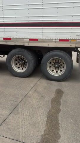 The video shows the side of a white semi-trailer with red and black stripes. The camera is focused on the trailer's tandem axles, which have four tires. The tires are black with silver rims. The trailer is parked on a concrete surface, which appears wet in places, possibly from recent rain or a spill. A red mud flap with a black logo is visible behind the rear tires. The video is from a low angle, looking up at the trailer.