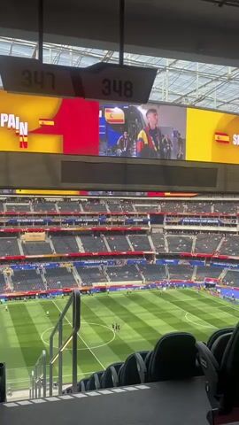 An aerial view of a large stadium from the perspective of a spectator in the upper seating section. The field below is a green soccer pitch with white markings, and several players are gathered in the center. Above the field, a large video screen displays the word "SPAIN" and the Spanish flag. The screen also shows clips of people in what appears to be a backstage area, possibly related to a sporting event. The seating in the stadium is arranged in multiple tiers, with many seats occupied by spectators. Two illuminated signs labeled "347" and "348" are visible hanging above the screen. The stadium has a modern, covered roof structure.