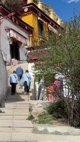 A low-angle shot shows four people walking up a stone staircase toward a traditional building. The people are dressed in casual clothes and are walking away from the camera. The building is several stories high and has a distinct architectural style, with white and yellow walls, red trim, and golden ornaments on the roof. The staircase is flanked by a stone wall on one side and a small tree on the other. The sky is blue with a few scattered clouds. The camera angle is slightly tilted upwards.