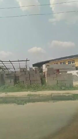 The video shows a view from a moving vehicle passing by a building. Initially, there is a utility pole and shipping containers next to a building with a beige exterior and many windows. As the camera moves, the building comes into full view, showing a curved, glass-paneled facade and a black roof. A fence runs along the front of the property, and there are piles of materials like concrete blocks and gravel nearby. The sky is visible with light cloud cover.