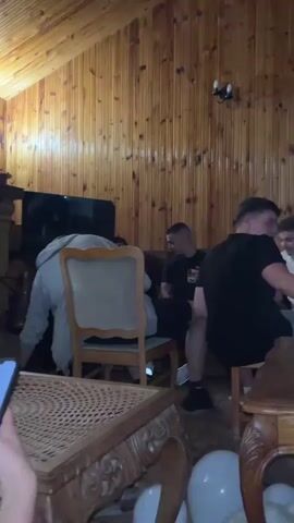 The video shows a group of men in a wood-paneled room, likely a cabin or lodge. One man is in a grey hoodie and is seated and gesturing with his arms. Another man is in a black shirt and is seated, holding a drink. A third man is in a black shirt and is standing. In the foreground, a person is holding a smartphone, looking at the screen. The coffee table in front of them is elaborately carved and cluttered with items, including beer bottles and what appears to be a white balloon.  The room has a rustic feel, with a fireplace, TV, and wood paneling on the walls and ceiling.  The lighting is warm and the focus is on the group and their interactions.