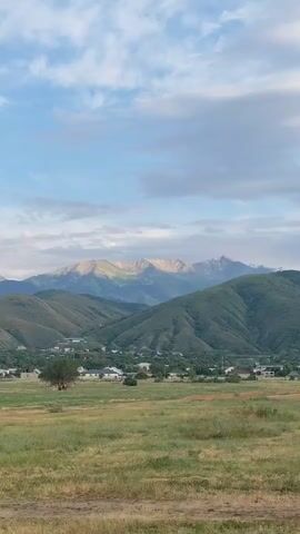 The media file shows a scenic, rural landscape with rolling green hills leading up to majestic, snow-capped mountains in the distance. A small village or town is nestled in the valley between the hills and mountains, with houses and trees visible. The sky is partly cloudy, with patches of blue sky peeking through. The lighting suggests it is either early morning or late afternoon, with the sun casting a warm glow on the mountain peaks. The foreground is a grassy field, appearing somewhat dry and sparsely vegetated, hinting at a possibly arid or semi-arid climate. There are no people or specific objects that draw significant attention, the focus being on the natural grandeur of the location. The overall mood is peaceful and serene, evoking a sense of natural beauty and tranquility. The location is identified as Қырғауылды, Kazakhstan. There is no visible text in the media file.