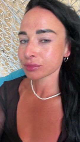 A woman with long black hair, freckles, a pearl necklace, and silver hoop earrings takes a drag from a small pink device and exhales vapor. She's wearing a sheer black top tied in the front, exposing some cleavage. She is sitting in what appears to be a wicker chair with a blue cushion. The background is blurry.