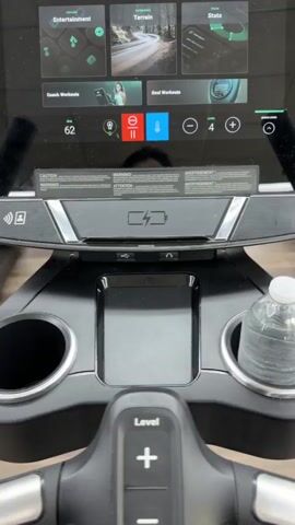 The video pans across a gym. In the foreground, a water bottle is in a cup holder on a piece of exercise equipment. In the background, several people are using ellipticals and treadmills. The gym has a wood-like floor and grey walls.