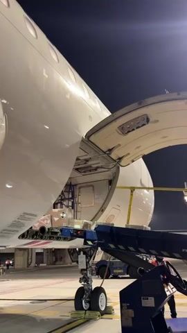 The video shows an airplane on the tarmac at night. A cargo loader is positioned next to the opened cargo bay of the aircraft, with boxes visible inside. The airplane's landing gear is visible. The camera pans across the side of the airplane, showing its white fuselage and windows. The video ends with a shot looking up at the underside of the airplane and its landing gear.