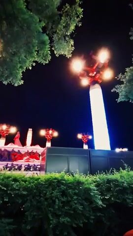 The video shows a night scene featuring a tall, illuminated column topped with a red structure supporting several lights. The base of the column emits a blue glow. In the background, there are other red-topped structures with lights, partially obscured by a gray wall. In the foreground, a hedge of green bushes is visible, with tree branches framing the top of the view. The sky is dark.