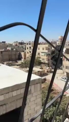 The video shows a view of a city from behind a black metal window grate. The buildings are a mix of tan and white, with flat rooftops. A street runs through the middle of the frame, with a few cars and pedestrians visible. The sky is blue and clear. There is a potted plant to the right of the window grate.