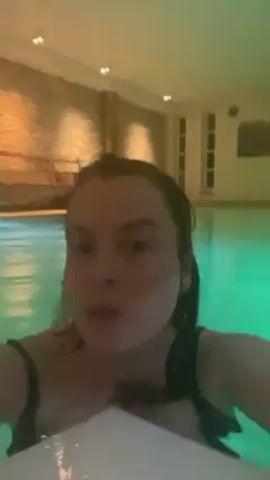A woman is in a pool, likely indoors, as suggested by the tiled wall behind her and the indirect lighting. She is wearing a black swimsuit with thin straps. Her hair is wet and appears to be pulled back. The pool is lit with green and possibly yellow or orange lights, giving the water a slightly greenish hue. She is looking directly at the camera, holding a white object in front of her, possibly a towel or a flotation device.