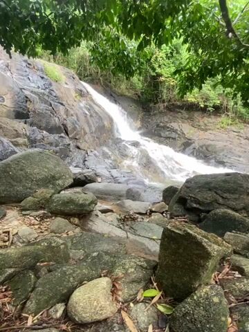 The video shows a rocky stream bed below a small waterfall. Water cascades down a sloping, rocky surface, forming the waterfall.  A woman is seen briefly at the top of the waterfall, then again near the base, wearing white and possibly taking a photo.  The streambed is filled with large, gray rocks, covered with some moss.  Green trees surround the waterfall.  The camera moves slightly, revealing more of the trees.