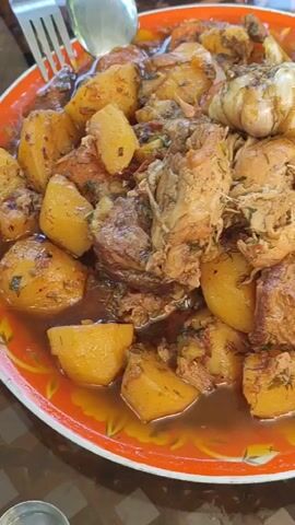 The video shows a close-up, overhead view of a large platter of food. The platter is orange with a decorative rim. The food appears to be a stew with large chunks of potato, meat, and what looks like a whole head of garlic on top. The ingredients are coated in a brown gravy and sprinkled with chopped green herbs. A fork and spoon are visible at the top left of the platter, suggesting the food is about to be served or eaten. The surface the platter rests on appears to be a table with a patterned tablecloth. A small silver bowl is partially visible in the lower left corner of the frame. The lighting suggests an outdoor or well-lit indoor setting.