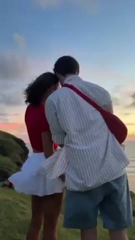 Here's a description of the video:

A young couple is standing on a grassy hill overlooking the ocean at sunset. The woman is wearing a red top and a white ruffled skirt, her hair blowing in the wind. The man is wearing a striped shirt, blue shorts, and a red crossbody bag. He turns and looks at the camera while holding his phone. The sky is a mix of blue, pink, and orange colors.