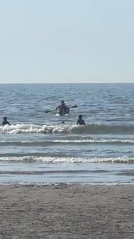 The video shows a beach scene with people in the ocean. In the foreground, a sandy beach is visible. The ocean is calm with small waves rolling onto the shore. Further out in the water, a group of people are present. Some are swimming and playing in the waves, while others are in a kayak or on a paddleboard. One person in the kayak appears to be paddling. The sky is clear and blue.