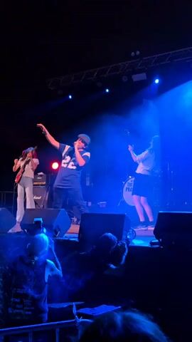 The video captures a band performing on a stage at night. A lead singer, wearing a baseball cap and a dark blue jersey with the number 22, is in the center, gesturing with his hands. To his left, a guitarist in a white shirt and light-colored pants plays a red electric guitar. To the singer's right, another guitarist, in a white shirt and dark shorts, plays an electric guitar, and behind them is a drummer with a drum kit that includes a bass drum with "THE" printed on it. The stage is lit by blue and red spotlights, with smoke effects adding to the atmosphere. The foreground shows the heads and shoulders of audience members, some holding up phones to record the performance. The stage is equipped with speakers and lighting rigs.