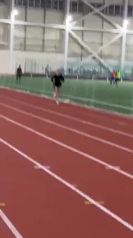 A person is running on an indoor track, wearing a black long-sleeved shirt and running shorts. The track is red with white lane lines and some small yellow markers. There is an artificial grass field beyond the track, where other people are visible. The building has a high ceiling supported by large white metal beams. There are lights overhead. The camera seems to be filming from a stationary position along the side of the track and then pans to follow the runner.