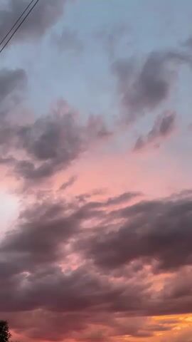 This video displays a time-lapse of a sunset with clouds. The sky transitions from a light blue at the top to shades of pink, orange, and red near the horizon. Darker, textured clouds are interspersed with lighter, wispy ones, creating a dramatic visual effect as the light changes. The lower portion of the video shows the faint silhouettes of trees or buildings against the vibrant sky, suggesting an outdoor, possibly rural or suburban, setting. There are no people or discernible actions within the frames.