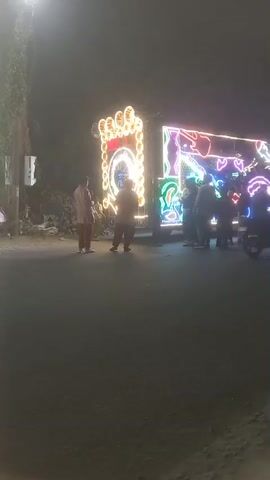 The video depicts a group of individuals standing around a brightly illuminated structure, possibly a float or vehicle decorated with neon lights. The scene takes place at night on a paved road, with the light from the structure being the most dominant feature. Several people are seen standing near the illuminated object, some are slightly obscured, while others are more clearly visible. Additionally, a scooter passes by in front of the people watching the decorated structure. Overall, the scene suggests a nighttime event with a mobile, illuminated display that has attracted an audience.