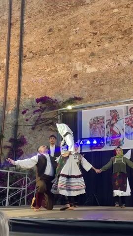 A group of people in traditional clothing are performing a dance on a stage in front of a stone wall. The dancers are dressed in colorful, embroidered outfits. The female dancer in the center is wearing a white headscarf, a white blouse, and a floral-patterned skirt. The male dancer in front of her is wearing a brown vest over a white shirt and brown pants. Other dancers are holding hands and moving in unison. There are lights on the stage, and a banner with images of people in traditional attire is displayed behind the dancers. A railing and some purple flowers are visible in the foreground and to the left of the stage. The overall scene suggests a cultural performance or festival.