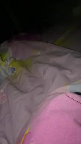 A woman with brown hair and blue eyes is lying down, possibly in a vehicle. She is wearing a light blue top and has a gold necklace. She looks up and smiles slightly, touching her hair. The background is blurry, showing what appears to be the interior of a van or similar vehicle. There is a pillow with a colorful design behind her head.