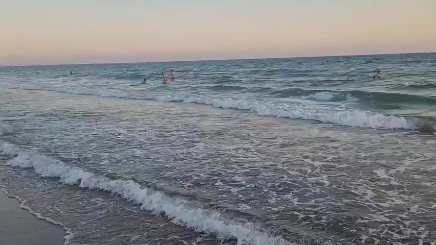 A wide shot of a beach shows gentle waves breaking on the shore. Several people are visible in the water, appearing to swim and play. The sky is a soft gradient of pink and blue, indicating either sunrise or sunset. The overall scene conveys a peaceful and relaxing atmosphere of a day at the beach.